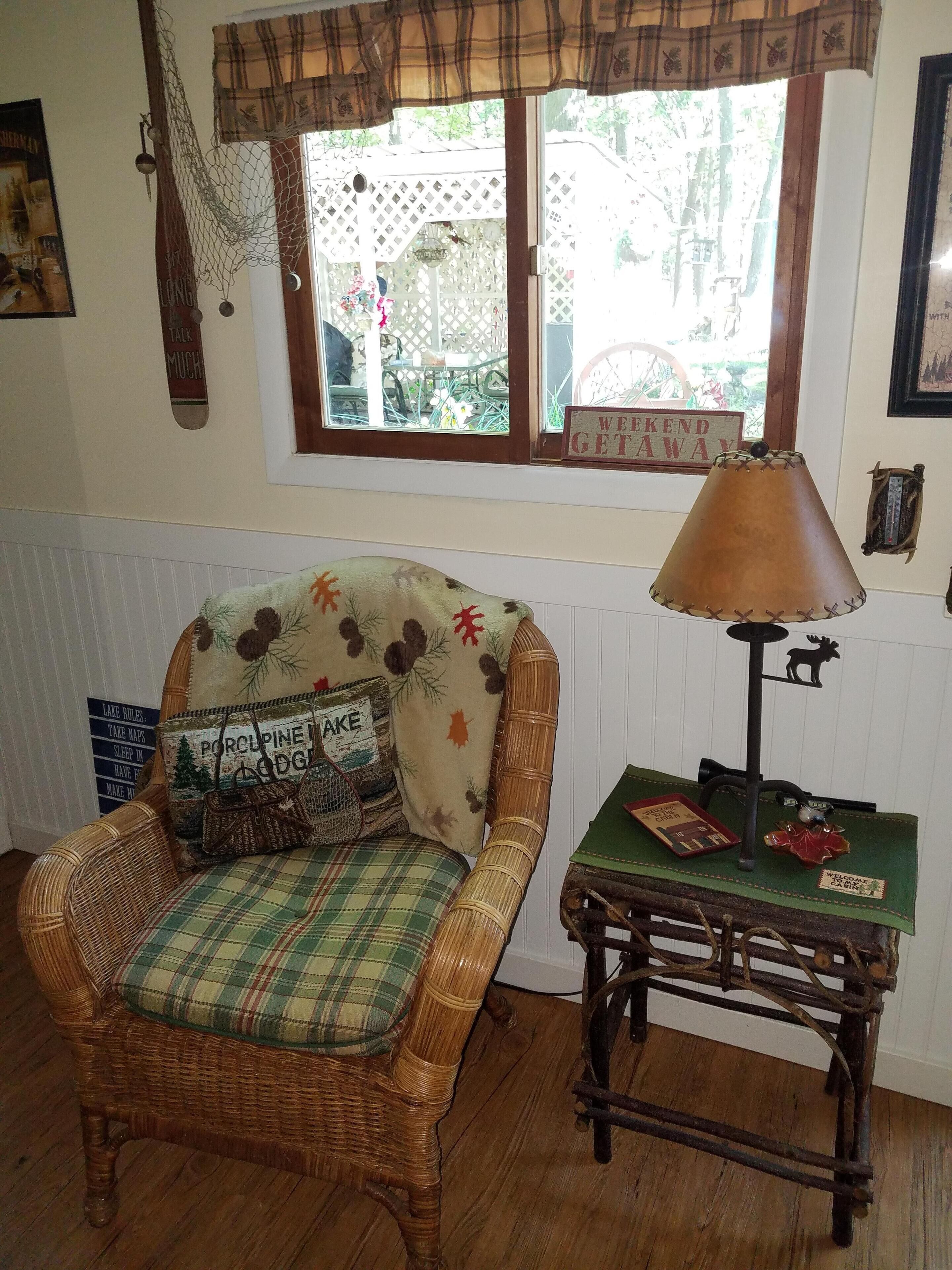 A wicker chair with a plaid cushion and a decorative pillow, a table with a lamp, and a window with a view of the outdoors.