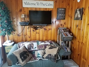 Interior d'una cabana amb una televisió, un arbre de Nadal, un sofà amb almofades decoratives i un cartell que diu "Welcome To The Cabin".