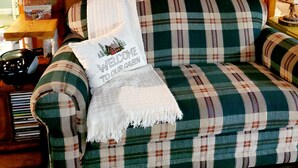 A cozy cabin interior with a plaid-patterned sofa, a welcome pillow, a lamp, and a wall-mounted sign reading "LAKE CABIN." -  Perfect getaway for couples ! close to Ludington!

 (Fountain)