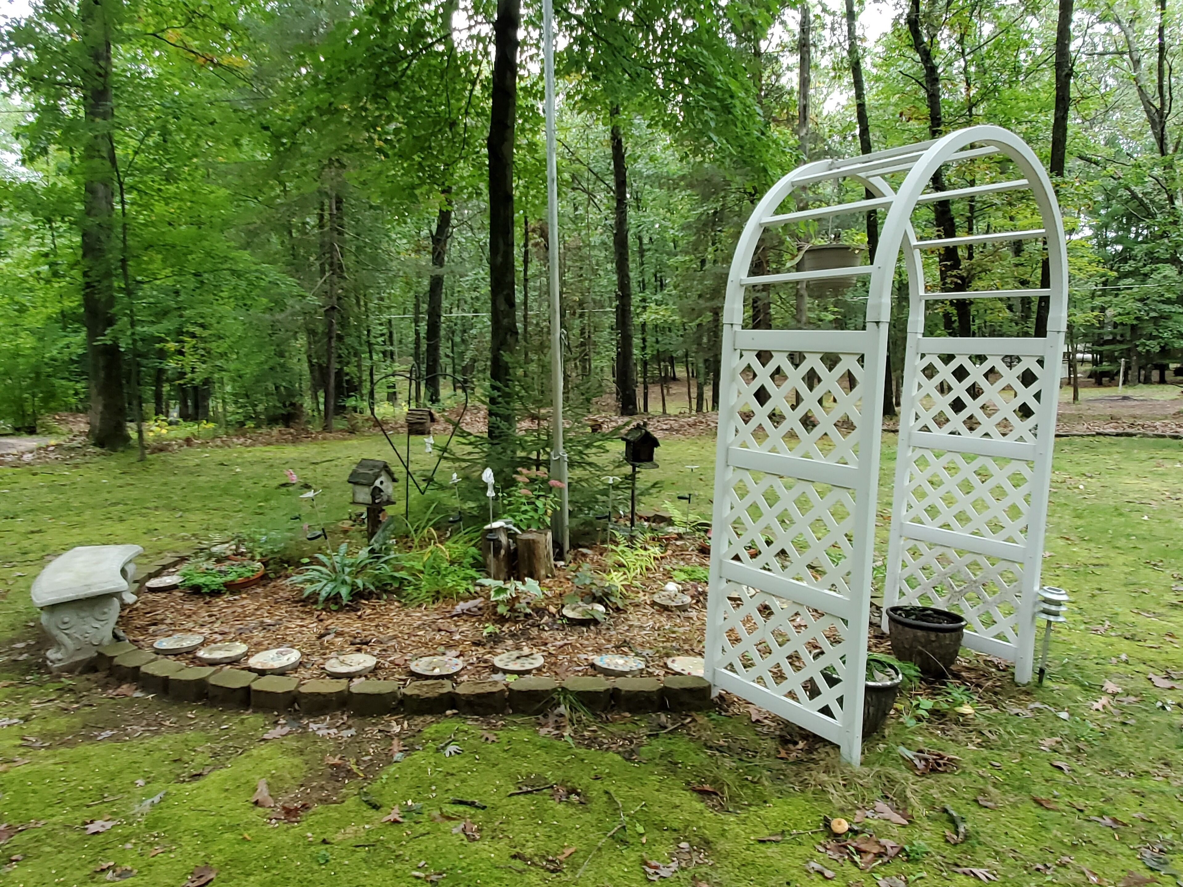Un jardín con un arco de celosía blanca rodeado de un sendero de piedra, comedores para pájaros y un nido de aves.