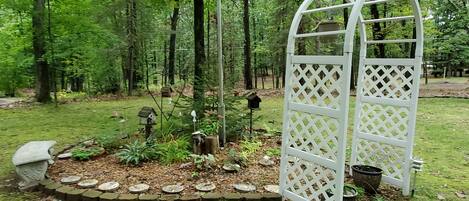 Un jardín con un arco de celosía blanca rodeado de un sendero de piedra, comedores para pájaros y un nido de aves.