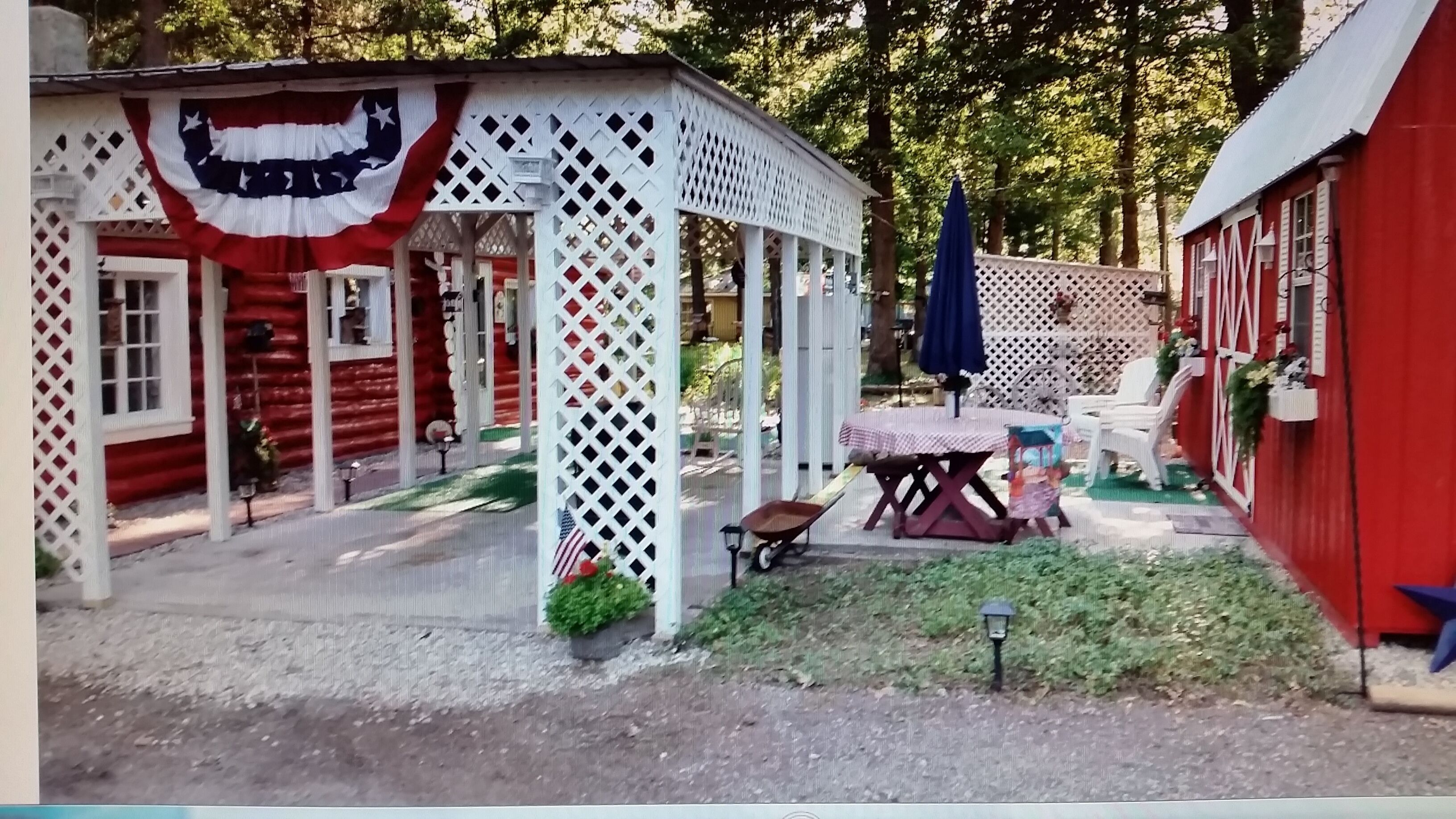 A rustic wooden cabin with a white lattice porch, a small table, and a wheelbarrow.