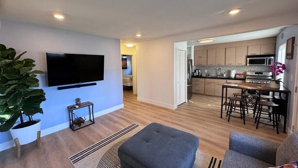 A modern living room with a flat-screen TV, a grey ottoman, and a sofa. A kitchen with wooden cabinets and a dining area with a table and chairs is visible in the background.