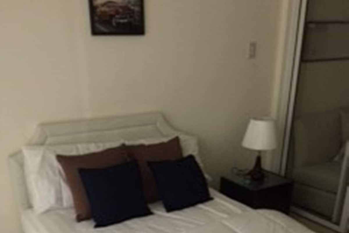 A neatly made bed with white linens and two dark pillows, a bedside table with a lamp, and a wall-mounted picture.