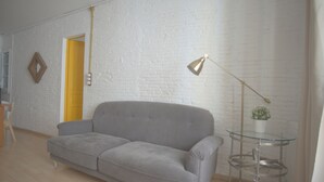 A living room with a grey sofa, a small glass table with a plant, a yellow door, and a white brick wall. - Charming flat next to the Cathedral (Girona)