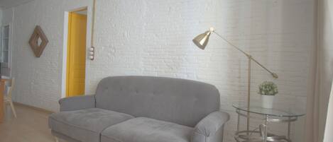 A living room with a grey sofa, a small glass table with a plant, a yellow door, and a white brick wall.