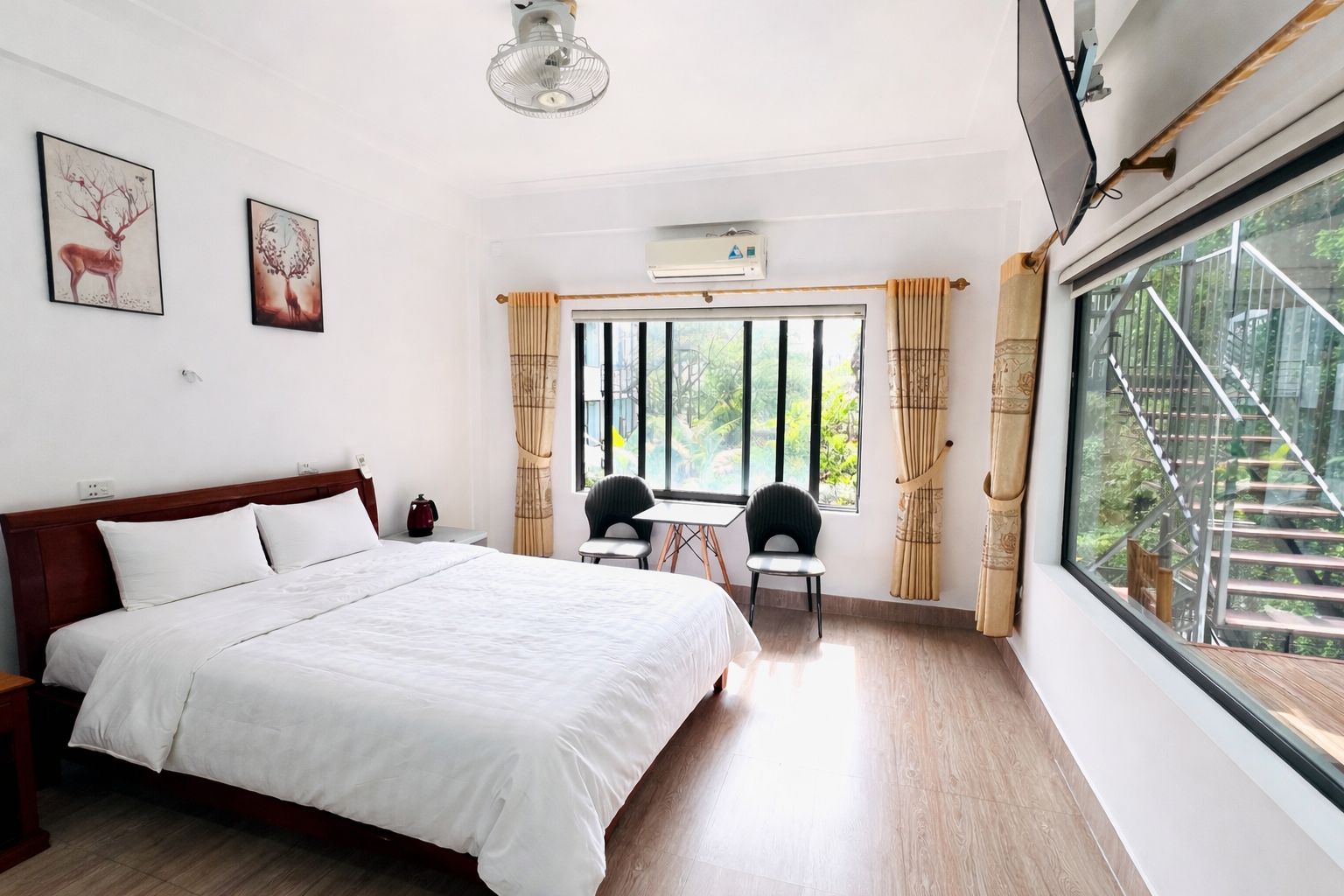 Habitación de hotel con una cama grande, un escritorio con dos sillas, una ventana con vista a follaje y obras de arte en las paredes.