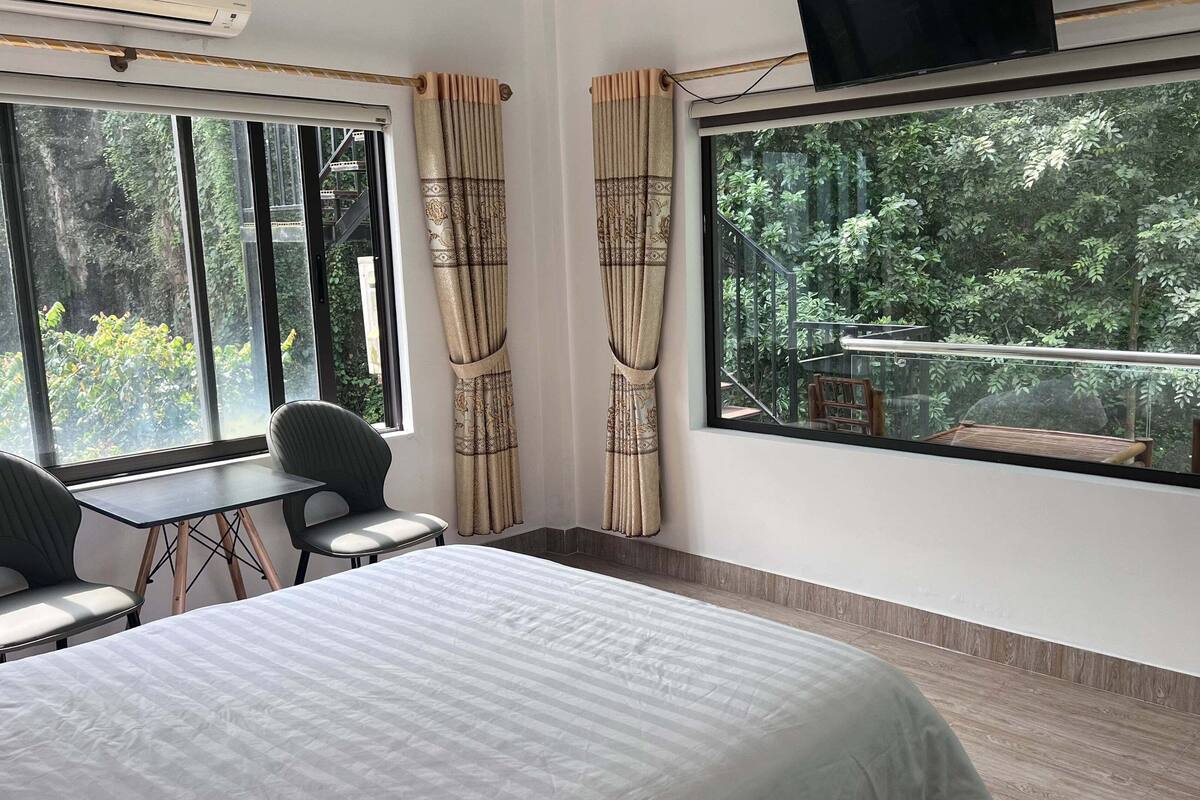 A hotel room with a bed, a desk, two chairs, a large window with a view of a forest, and a TV mounted on the wall.