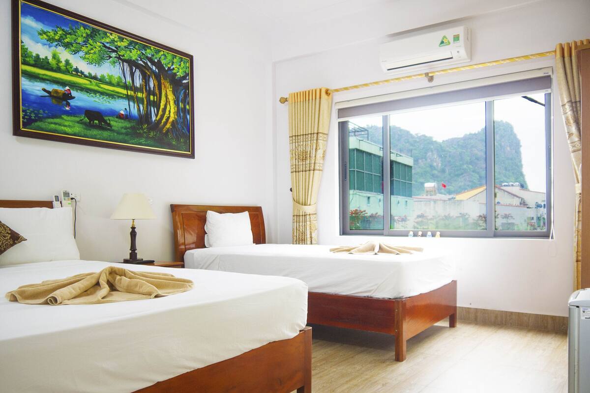 A hotel room with two beds, a painting of a landscape, an air conditioning unit, and a window with a view of buildings.