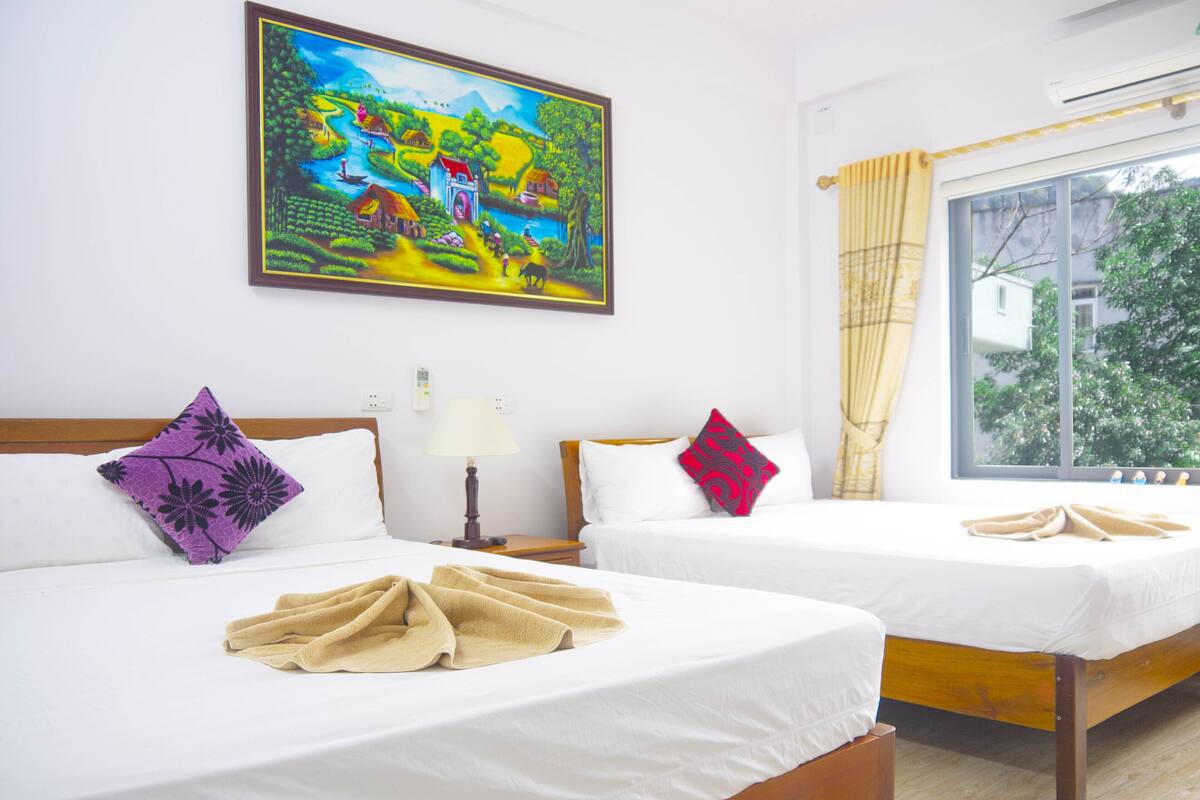 A hotel room with two beds, a painting on the wall, a window with curtains, and a bedside table with a lamp.