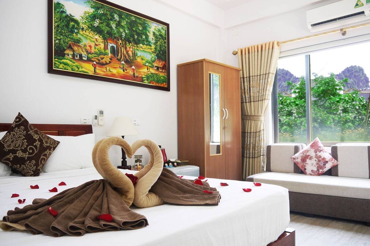 A hotel room with a bed, a painting, a window with a view, and a heart-shaped towel arrangement.