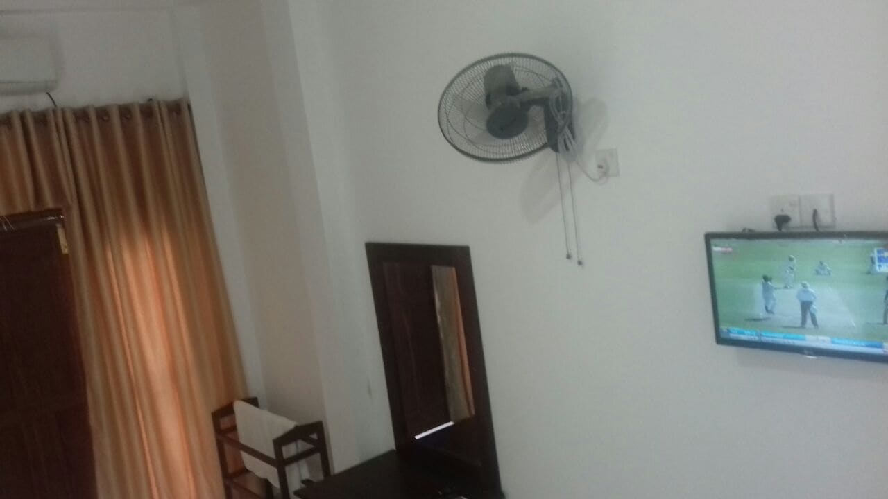 Habitació amb ventilador de sostre, televisió que mostra un partit esportiu, una porta de fusta i una cortina.