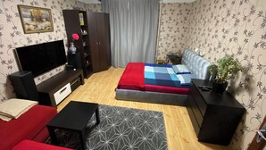 Een knus woongedeelte met een bank, televisie, bed met een beddengoed met patroon, een klein tafeltje, een boekenkast en een potplant.