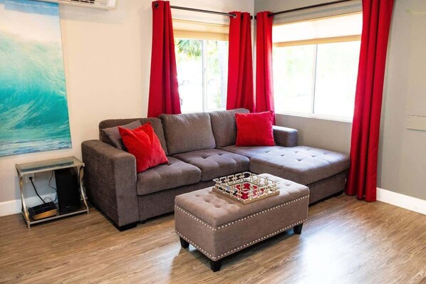 A living room with a grey sectional sofa, red cushions, a wooden coffee table, and a large window with red curtains.