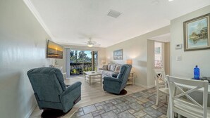 A spacious living room with two armchairs, a sofa, and a dining area. A balcony with a view of greenery is visible through the glass doors. - Condo # 110 Ground floor unit with quick access to Siesta Beach Amenities (Sarasota)
