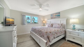 A bedroom with a bed, a ceiling fan, a window, a nightstand, a lamp, and a TV. - Condo # 110 Ground floor unit with quick access to Siesta Beach Amenities (Sarasota)