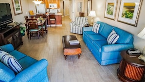 A living room with a blue sofa, a coffee table, and a TV. There is a dining area with wooden chairs and a table.