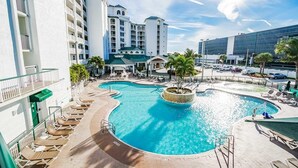 A hotel pool area with multiple lounge chairs, a central water feature, and a building with balconies.