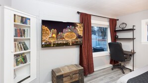 A room with a bookshelf, a desk, a chair, a window with curtains, and a large TV displaying a cityscape. - The Immaculate Guest Cottage In Downtown Coeur  D'Alene (Coeur d'Alene)