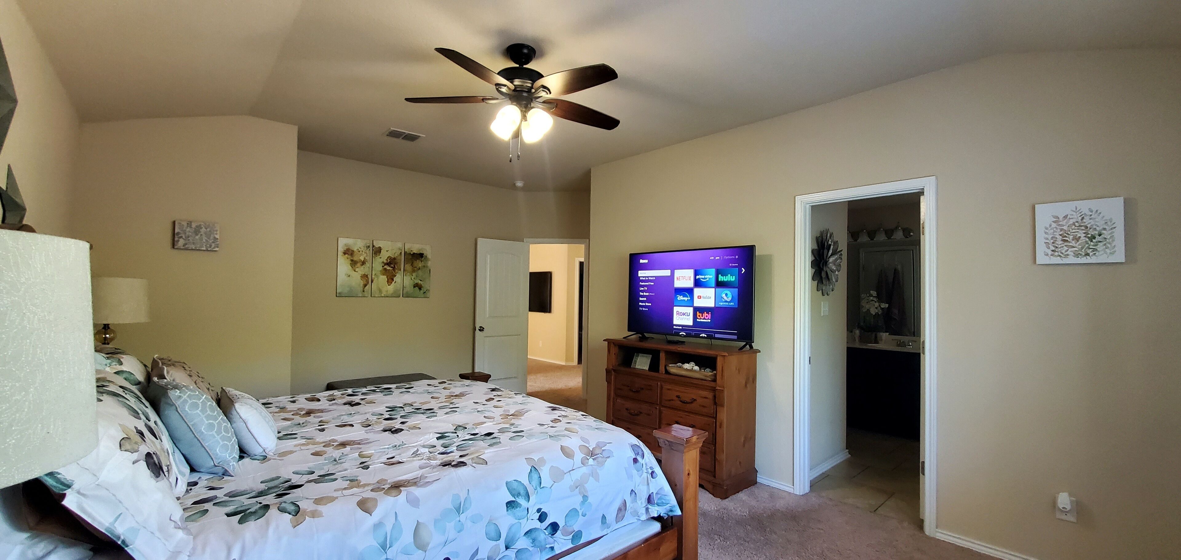 Un dormitorio con cama, ventilador de techo, televisor mostrando servicios de transmisión y una puerta que da a otra habitación.
