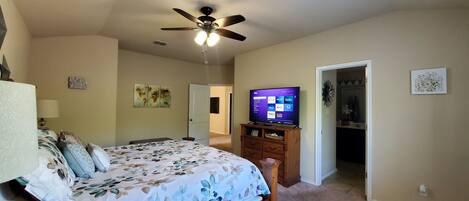 Un dormitorio con cama, ventilador de techo, televisor mostrando servicios de transmisión y una puerta que da a otra habitación.