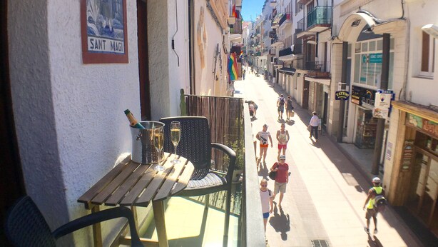 Uma rua estreita com edifícios em ambos os lados, uma mesa de madeira com cadeiras e uma garrafa de champanhe.