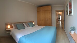 Quarto com cama grande, guarda-roupa de madeira, mesa de cabeceira com luminária e um vaso com planta.