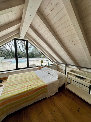 A bedroom with a sloped ceiling, wooden beams, a bed with a striped bedspread, a bedside table, and a view of an outdoor area with a table and chairs. - Dream location- Modern villa with pool by the sea, just a stone's throw from Rome. (Anzio)