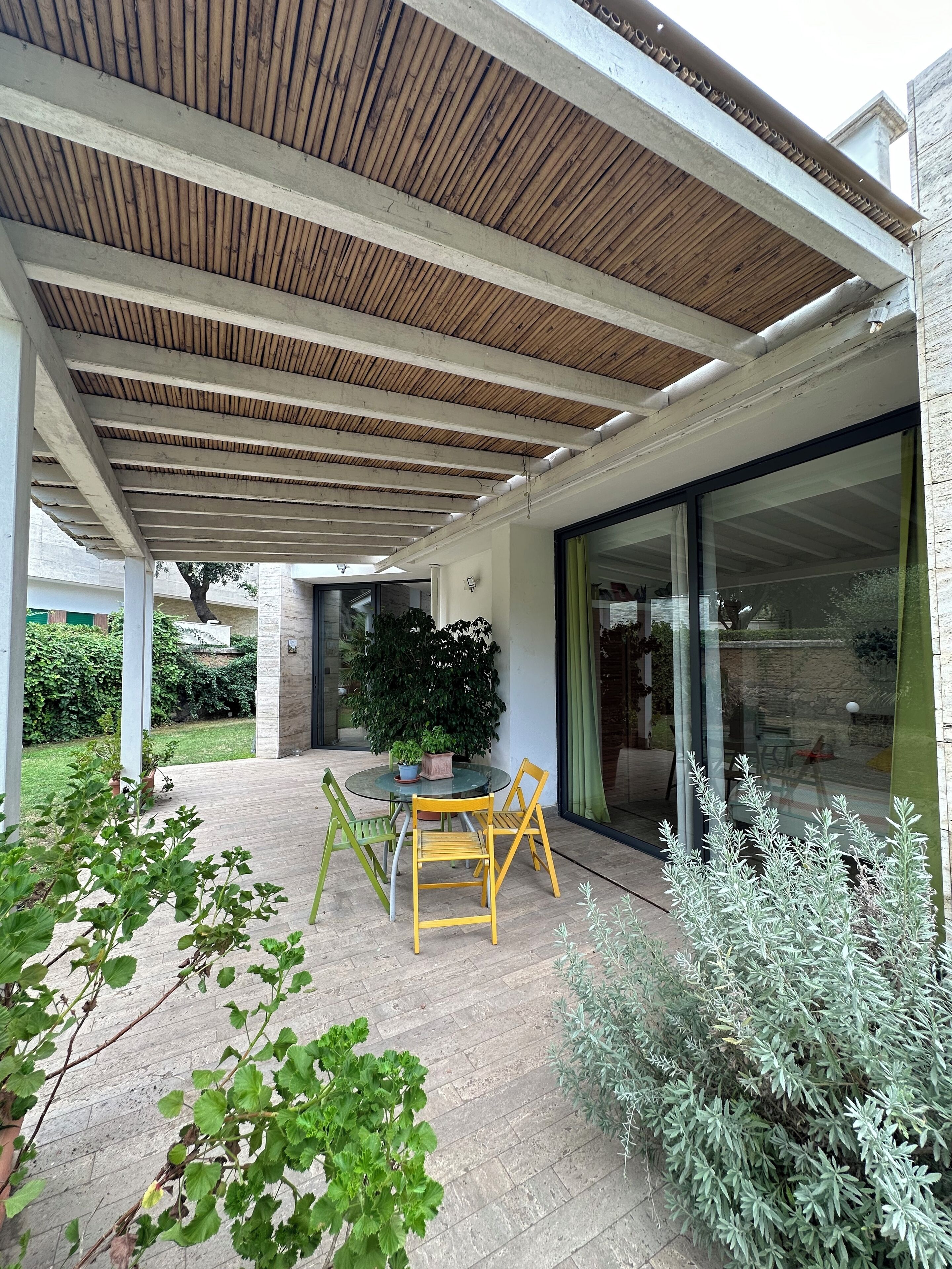 A covered patio area with a table and chairs, surrounded by greenery and a glass door leading to an interior space.