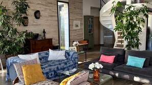 A modern living room with a sofa, armchairs, and a coffee table. There are plants, a bookshelf, and a spiral staircase.