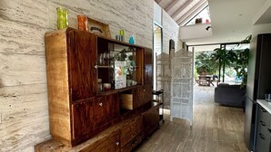 A spacious room with a wooden cabinet against a marble wall, a dining area with a table and chairs, and a view of the outdoors.