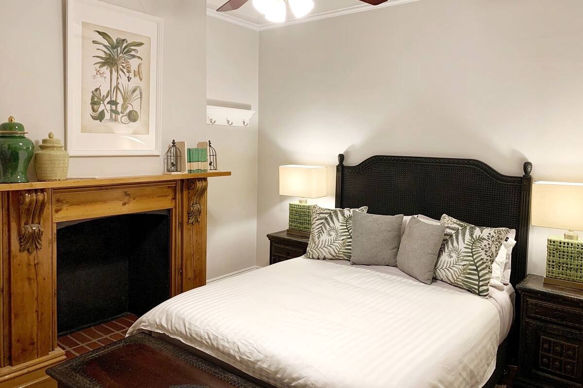 Quarto com cama, duas mesinhas de cabeceira com luminárias, lareira de madeira e ventilador de teto.