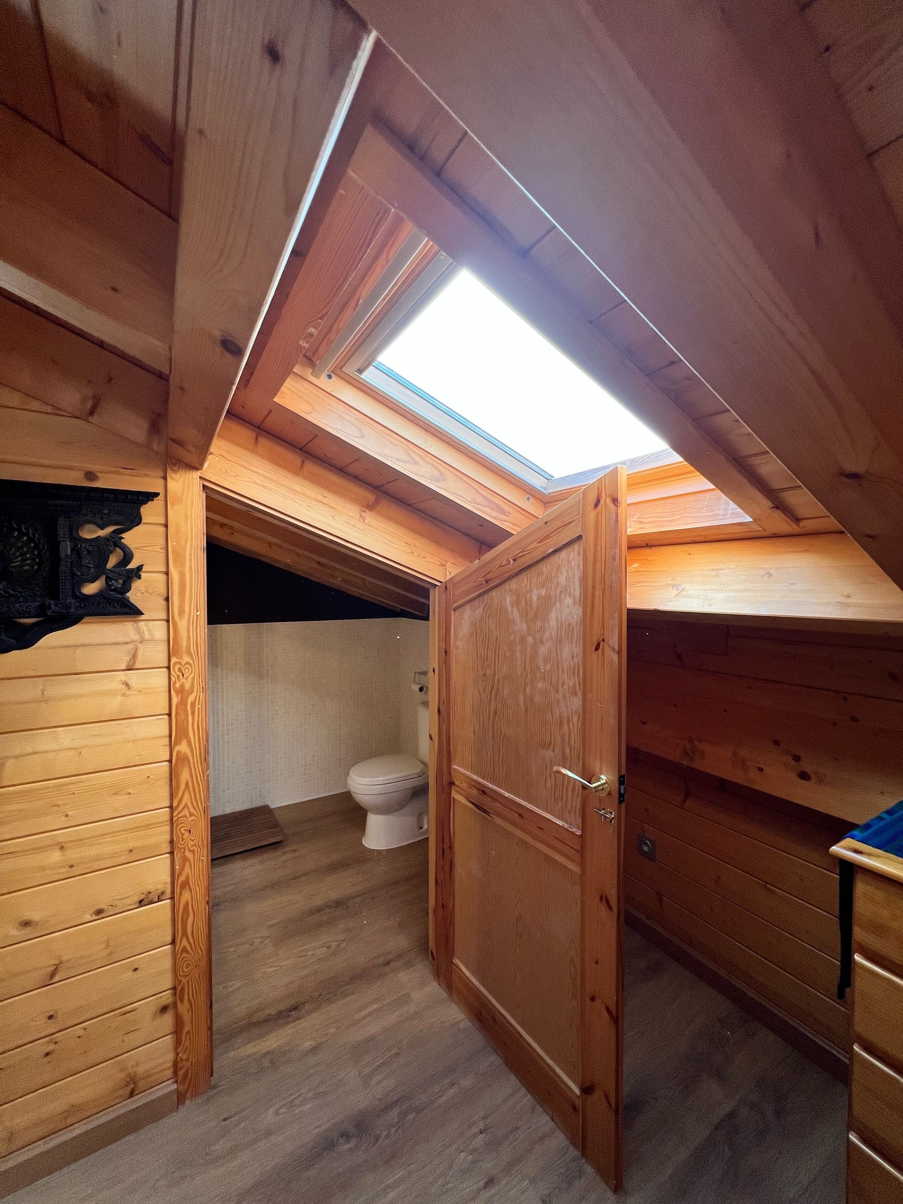 Une petite chambre mansardée en bois, avec un puits de lumière, des toilettes et une armoire en bois.