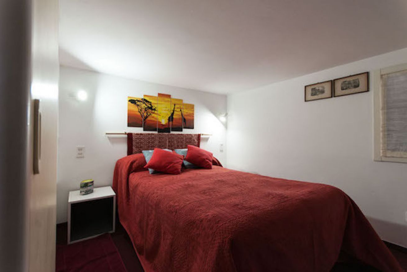 Un dormitorio con colcha roja, una mesita de noche con lámpara y un cuadro colgado en la pared.