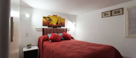 Un dormitorio con colcha roja, una mesita de noche con lámpara y un cuadro colgado en la pared.