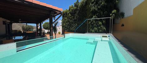 Una piscina con superficie de agua azul cristalina, rodeada por un área techada con macetas y plantas.
