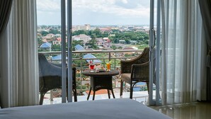 Un balcón con sillas de mimbre y una mesa, con vistas a un paisaje urbano con edificios y zonas verdes.