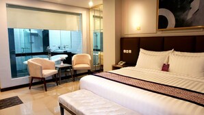 Habitación de hotel con una cama grande, un rincón de estar con dos sillones, una mesita, un baño con bañera y un espejo amplio.