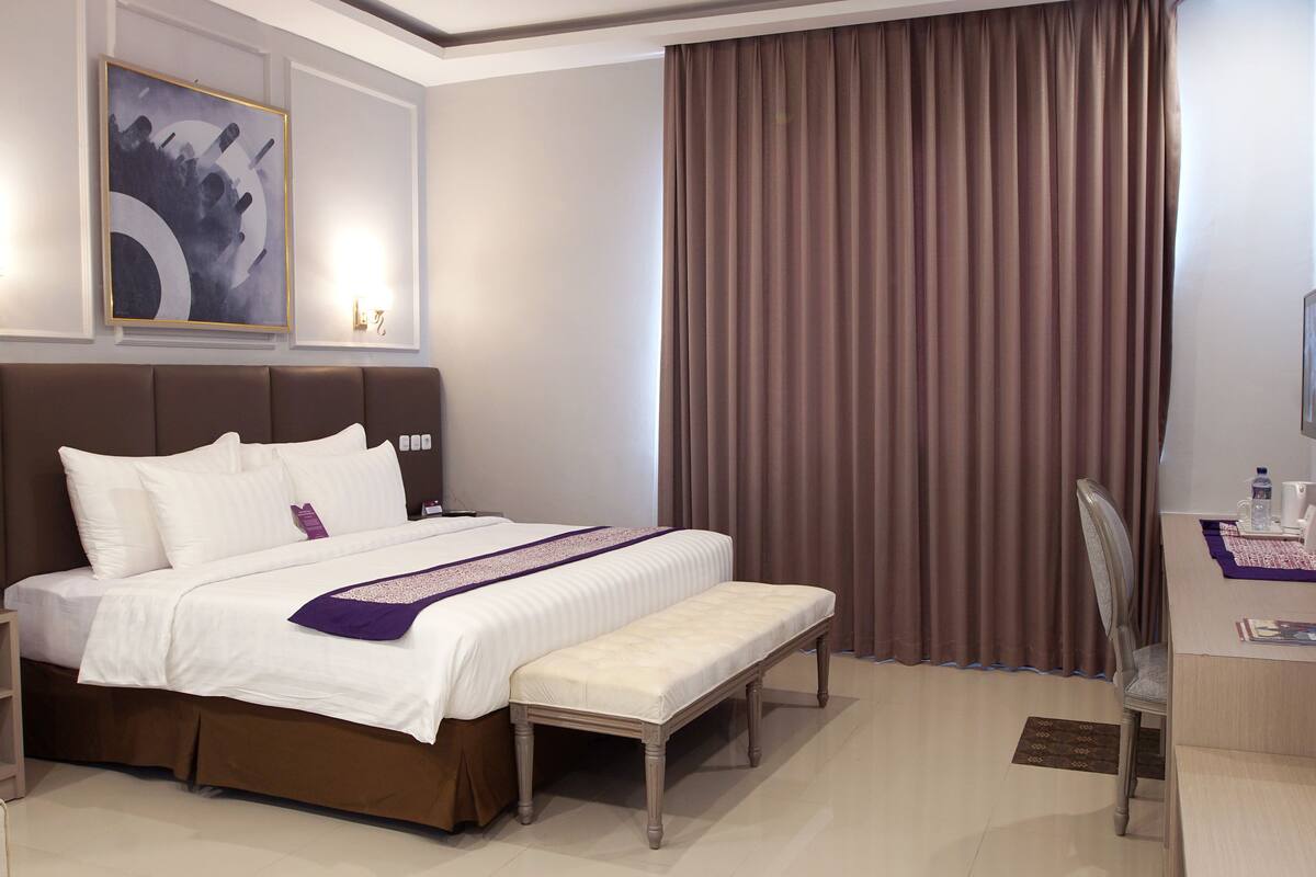 Habitación de hotel con una cama grande, un escritorio con silla, televisión y ventana con cortinas.