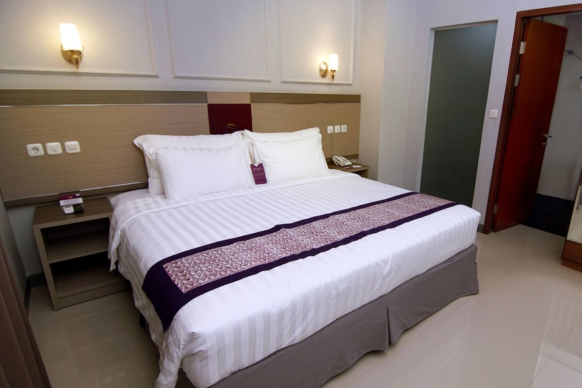 Habitación de hotel con una cama grande, una mesita de noche con teléfono y control remoto, un espejo y una puerta que da a otra habitación.