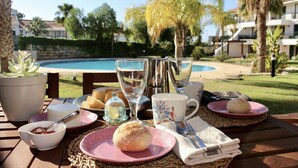 A table set with breakfast items including pastries, a cup, and a glass of water, with a pool and palm trees in the background. - Exclusive 2 bed  apartment in Vila Sol Village by Experience MamiMi (Quarteira)