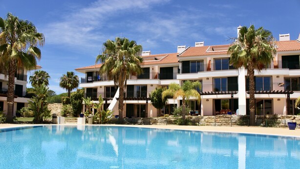 A swimming pool surrounded by multi-story buildings with balconies and palm trees. - Exclusive 2 bed  apartment in Vila Sol Village by Experience MamiMi (Quarteira)
