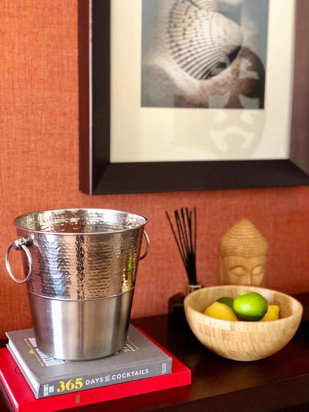 A metallic cup on a red book titled "365 Days of Cocktails," a wooden bowl with fruits, and a framed picture on a wall.