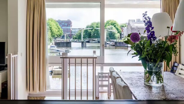 Een kamer met een groot raam dat uitkijkt op een gracht met boten, een tafel met een bloevas en een stoel.