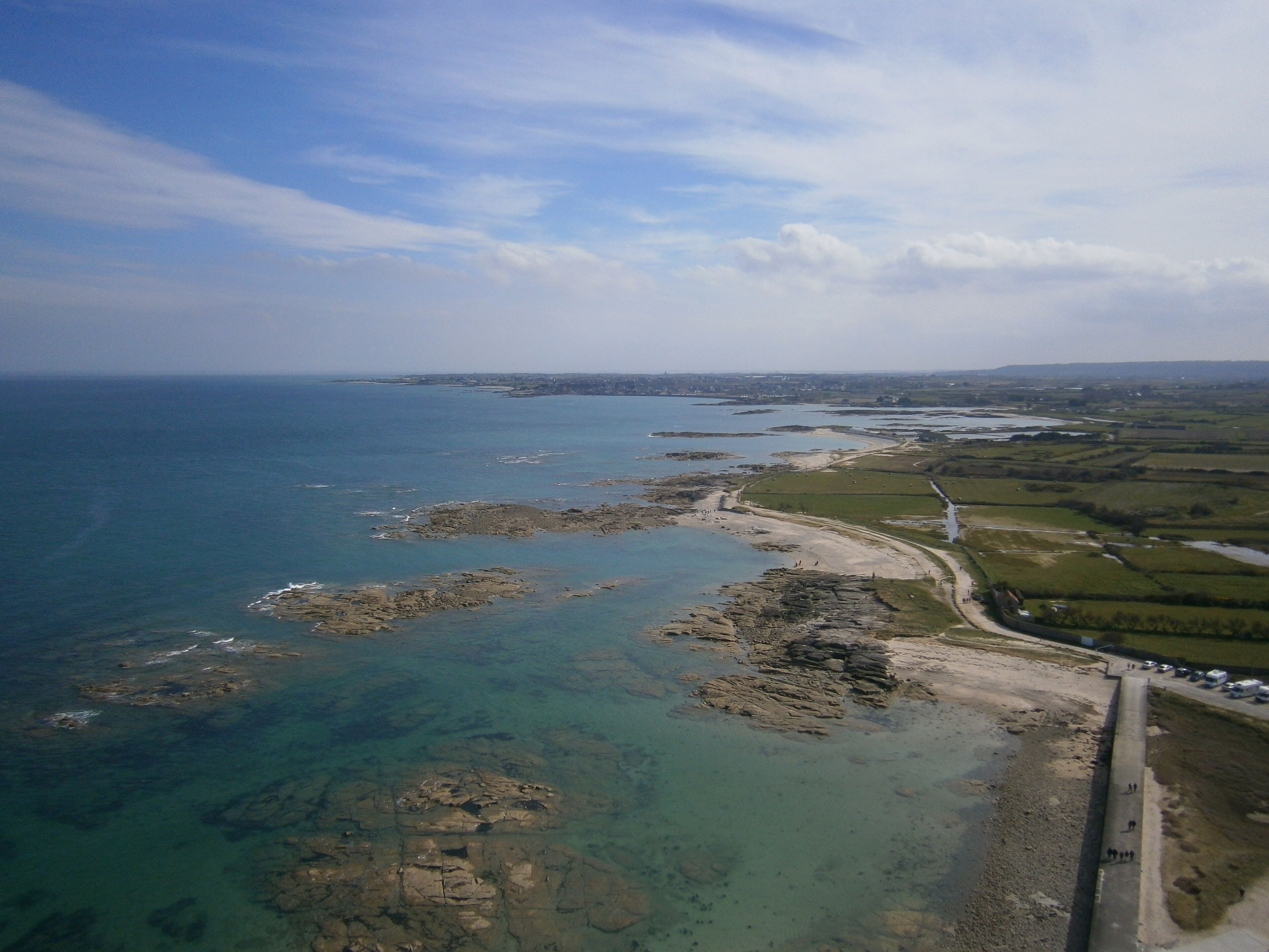 An aerial view of a coastal area with rocky outcrops, clear blue waters, and a paved path along the shore.