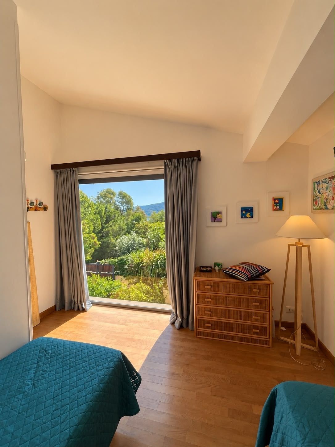 Una habitación con un ventanal grande, un aparador de madera, una lámpara de pie y dos camas con ropa de cama color turquesa.