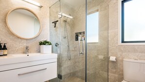 A modern bathroom with a glass-enclosed shower, a round mirror, a white sink, and a toilet. - Guest House on Queens (Waiheke Island)