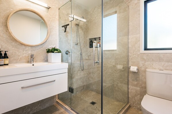 A modern bathroom with a glass-enclosed shower, a round mirror, a white sink, and a toilet. - Guest House on Queens (Waiheke Island)