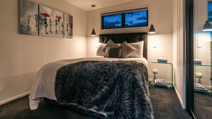A modern bedroom with a large bed, a headboard with multiple pillows, a glass bedside table, and a painting on the wall. - Guest House on Queens (Waiheke Island)