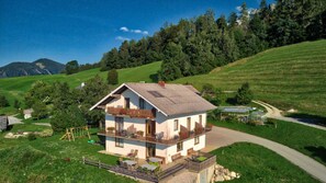 Ein zweigeschossiges Haus mit Balkon, eingebettet in eine ländliche Landschaft mit Hügeln und Bäumen.